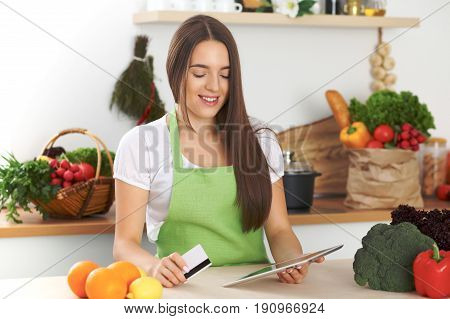 Young woman is making online shopping by tablet computer and credit card. Housewife found new recipe for cooking in a kitchen.