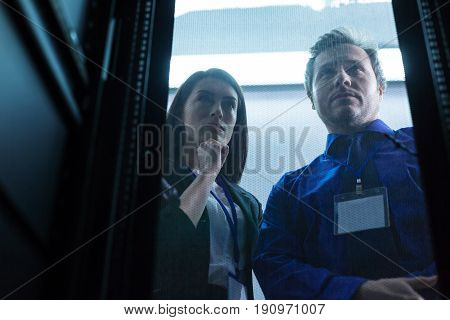 Technological problem. Pleasant thoughtful serious woman standing with her colleague and holding her chin while thinking about their technological problem