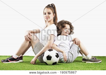 Girl and boy are sittng backs to each other on the grass with a soccer ball. Two children soccer players in sports smile over a white background
