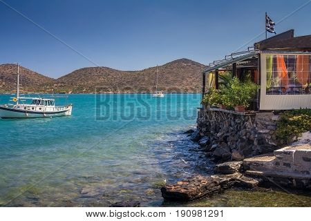 Cafe on the shore at the water's edge. Two boats are moored side by side. Low mountains in the background. Gulf of Mirabello. Elounda. Crete. Greece.