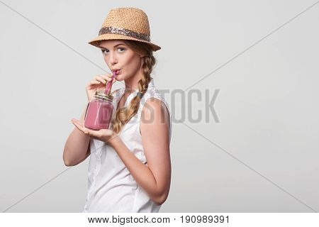 Smiling girl with jar tumbler mug drinking through straw a pink smoothie drink, portrait over grey background with blank copy space