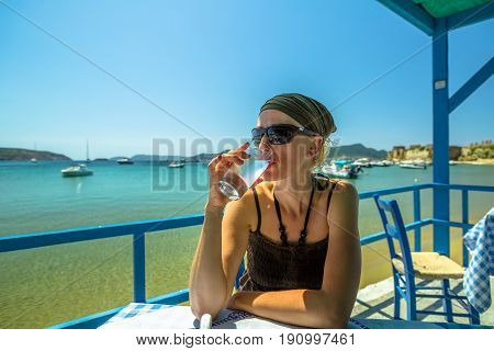 Woman drinking fresh water or lime juice at Methoni Beach. Female is looking away while sitting at an outdoor restaurant. Methoni Castle on background, Peloponnese, Greece. Europe summer vacation.