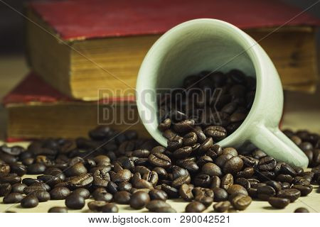 Coffee Bean In Tilted Ceramic Cup And Old Book Laid Behind On Wooden Table With Morning Light.