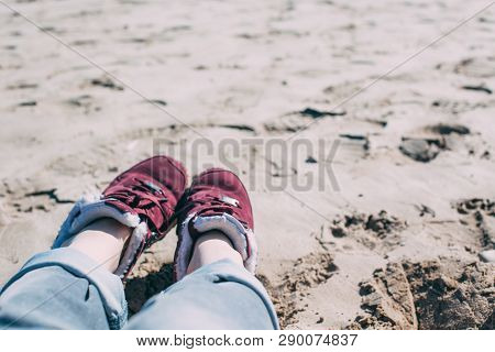 Feet Of Young Woman In Sport Footwear And Jeans Relaxing After Hiking On Sand Beach. Pov. Adventure 