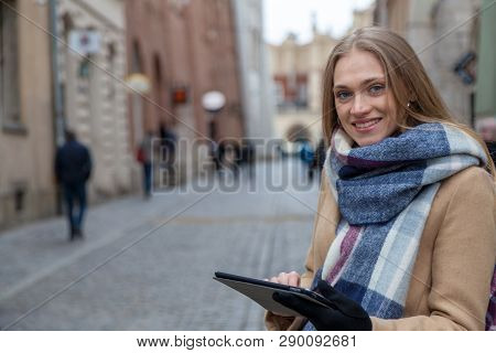 Blonde Beautiful Woman Holding Tablet On City Street. Portrait Of Young Woman Wearing Tartan Scarf S