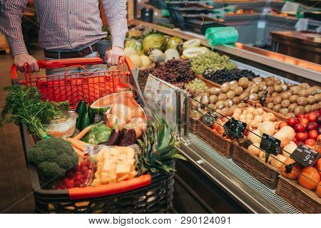 Young Man Alone In Grocery Store. Cut View Guy Hold Trolley Hand. Basket Full Of Vegetables And Frui