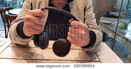 A Man Holds In Two Hands Black Modern Headphones Over Wooden Table In Outdoor Cafe