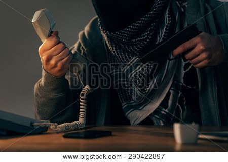 Cropped View Of Terrorist Holding Handset And Gun At Table