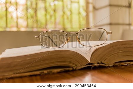 An Open Book And A Eyeglass Nicely Placed On Wooden Table On A Warm Sunny Morning, Side View Close U