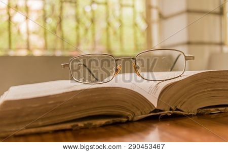 An Open Book And A Eyeglass Nicely Placed On Wooden Table On A Warm Sunny Morning, Side View Close U