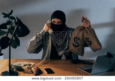 Angry Terrorist Holding Money Bags With Dollar Signs And Talking On Telephone