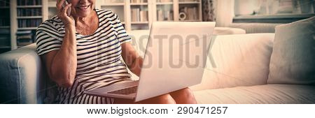 Senior woman talking on mobile phone while using laptop in living room at home