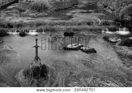 Long Exposure Of The River Flowing Past A Sword Sticking Out Of The River In  Cheddar Village In Som
