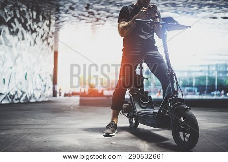 Bearded Tattooed Male Hipster Using Mobile Phone After Riding By Electric Scooter In The City.