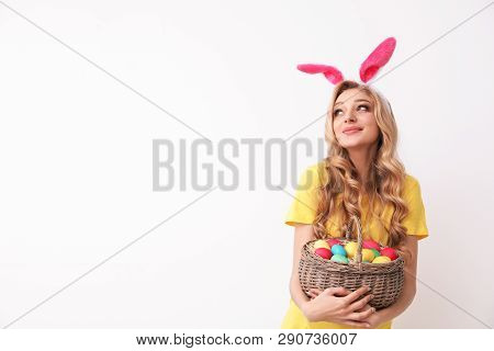 Beautiful Young Woman In Bunny Ears Headband Holding Basket With Easter Eggs On Light Background, Sp