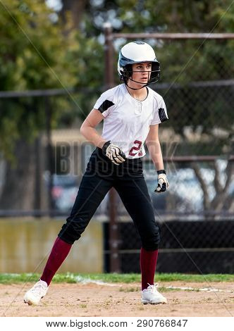 Female Teen Softball Player In Black And White Uniform Taking A Cautious Lead Off From First Base.