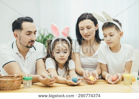 Inspired Family In White T-shirts Sitting Together At The Table, Holding Colored Easter Eggs In Thei