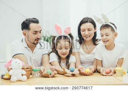 Inspired Family In White T-shirts Sitting Together At The Table, Holding Colored Easter Eggs In Thei
