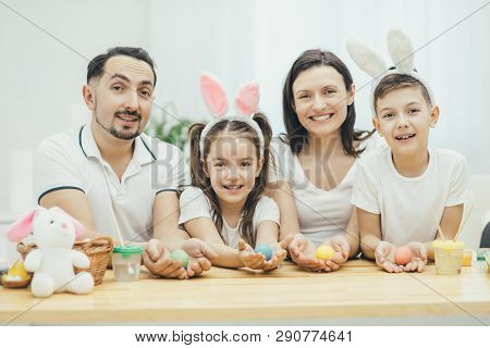 Inspired Family In White T-shirts Sitting Together At The Table, Holding Colored Easter Eggs In Thei