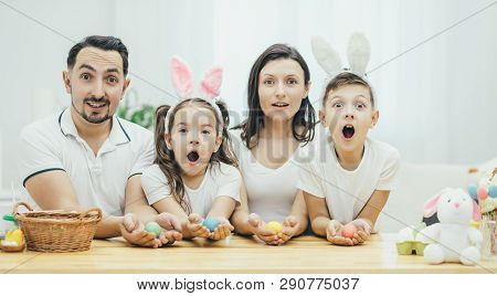 Inspired Family In White T-shirts Sitting Together At The Table, Holding Colored Easter Eggs In Thei