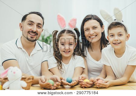 Inspired Family In White T-shirts Sitting Together At The Table, Holding Colored Easter Eggs In Thei