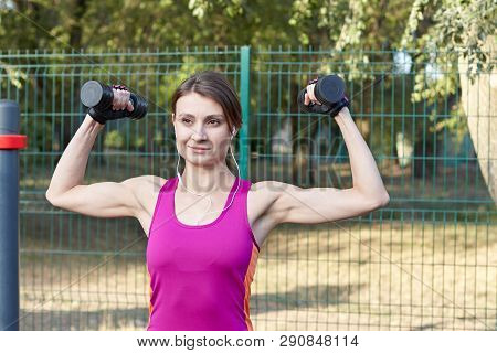 Young Slim And Sporty Woman In Bright Sportswear Trains With Dumbbells For Biceps At Outdoor Sportgr