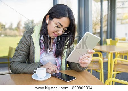 Smiling Focused Girl Transferring Data From Tablet To Smartphone. Young Woman In Earphones Setting P