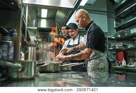 Extreme Cooking. Profesional Chef Teaching His Two Young Trainees How How To Flambe Food Safely. Res