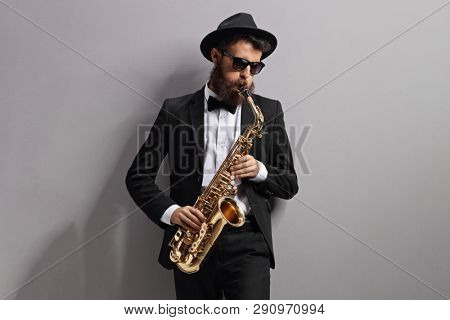 Man playing a sax and leaning against a gray wall