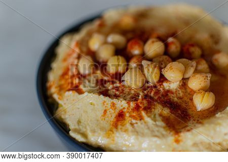 Сreamy Hummus With Chickpeas And Red Yellow Spices In Dark Plate In Light Kitchen. Vegetarian Food. 