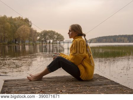 A Barefoot Girl With Her Hair In Braids Sits On A Wooden Bridge By The Lake Against The Sunset. The 