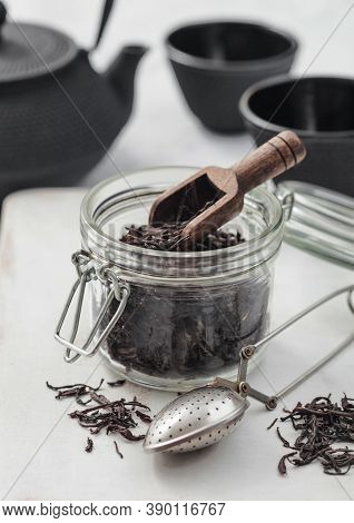 Black Loose Organic Tea In Glass Jar With Scoop And Strainer Infusor On Light Table Background With 