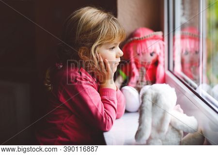 Cute Toddler Girl Sitting By Window And Looking Out On Rainy Day. Dreaming Child With Doll And Soft 