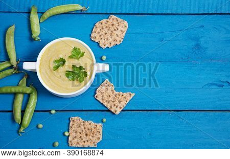 Pea Soup-puree In A White Cup On A Blue Wooden Background, Next To Pea Pods And Grain Crackers. Plac