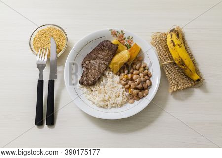 Plate Of Rice, Beans, Steak And Cassava. With Cutlery And Manioc Flour And Banana For Dessert. White