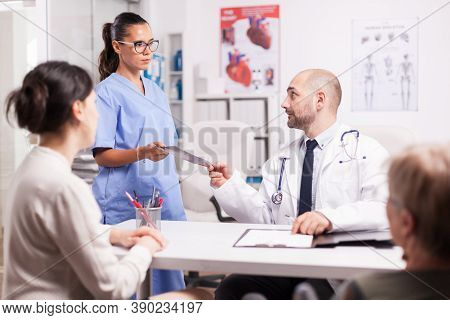 Nurse Giving Doctor Patient X-ray During Examination In Hospital Office. Daughter And Her Disabled M