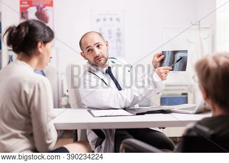 Doctor Showing Knee Injury On X-ray Image In Hospital Office To Young Woman And Her Disabled Mother 