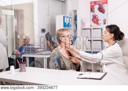 Young Female Doctor Feeling Senior Patient Neck For Swollen Glands In Hospital Cabinet Wearing White
