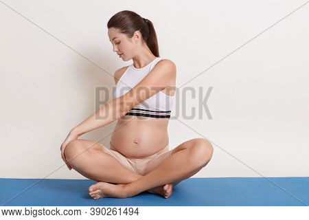 Beautiful Pregnant Woman Doing Yoga Exercises, Expectant Mother Wearing White Bra And Beige Leggins 