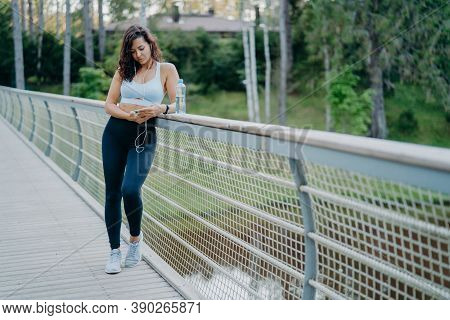 Sporty Slim European Woman In Active Wear Checks Notification On Mobile Phone, Listens Music From Pl