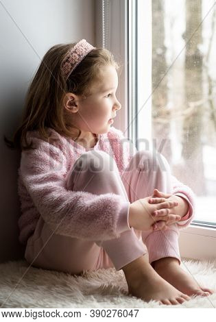 Kid Sitting On Windowsill At Home Looks Out Window To Snow, Portrait Of Pretty Little Girl On Fur Ru