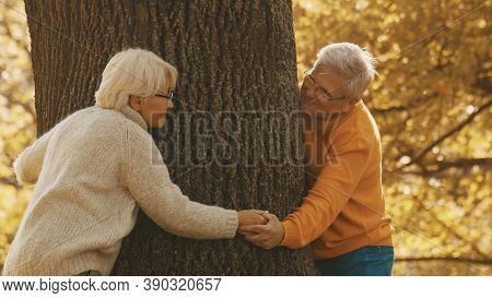 Old Couple Having Romantic Autumn Day In Forest. Hugging Tree And Smiling. High Quality Photo