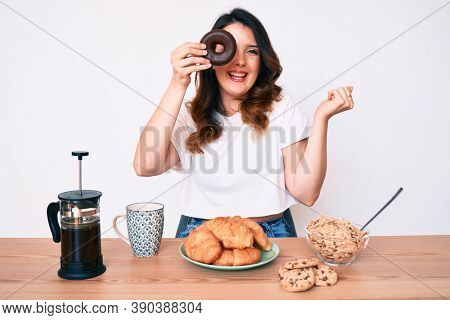 Young beautiful brunette woman eating breakfast holding cholate donut screaming proud, celebrating victory and success very excited with raised arm 