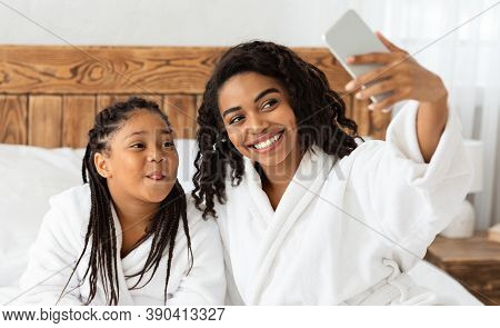 Selfie Fun. Cheerful African Mom And Daughter In White Bathrobes Taking Photo On Smartphone, Capturi