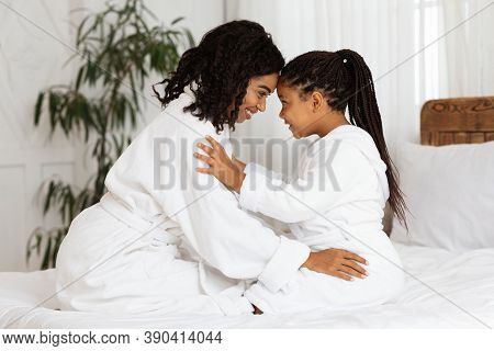 Loving Black Mother Bonding With Her Little Daughter On Bed At Home, Wearing Bathrobes, Relaxing Tog