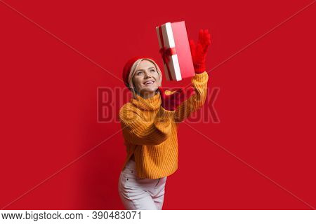 Smiling Caucasian Woman Is Wearing Gloves And Hat On A Red Wall While Catching A Present Box