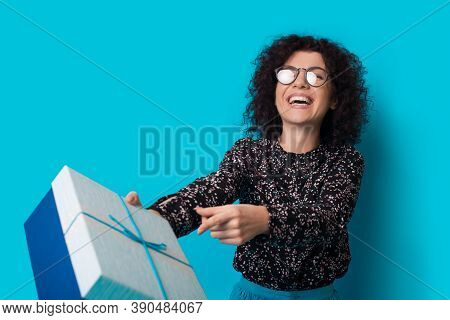 Lovely Curly Haired Woman Is Smiling At Camera And Receiving A Present On A Blue Studio Wall