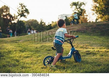 Lovely Caucasian Boy Riding The Bike In The Park Playing On The Green Grass And Looking At The Other