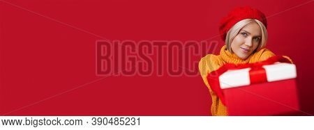 Cute Blonde Woman Wearing A Hat Is Showing At Camera A Present Posing On A Red Wall With Free Space