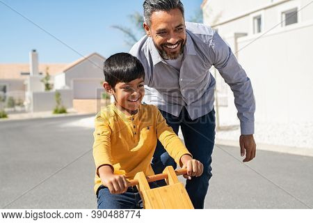 Cheerful middle eastern father helping excited son to ride wooden balance cycle on street. Happy boy enjoying riding bycicle with his dad. Dad teaching his indian son to ride bicycle outdoor.